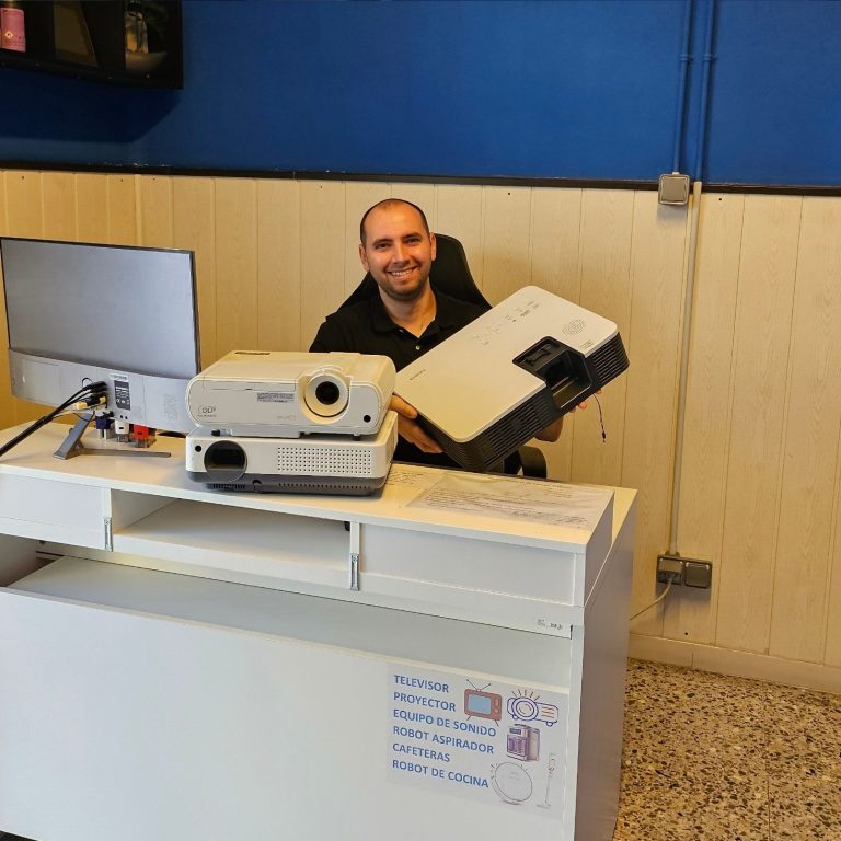 Hombre sonriente sentado frente a un escritorio, sosteniendo un proyector averiado.